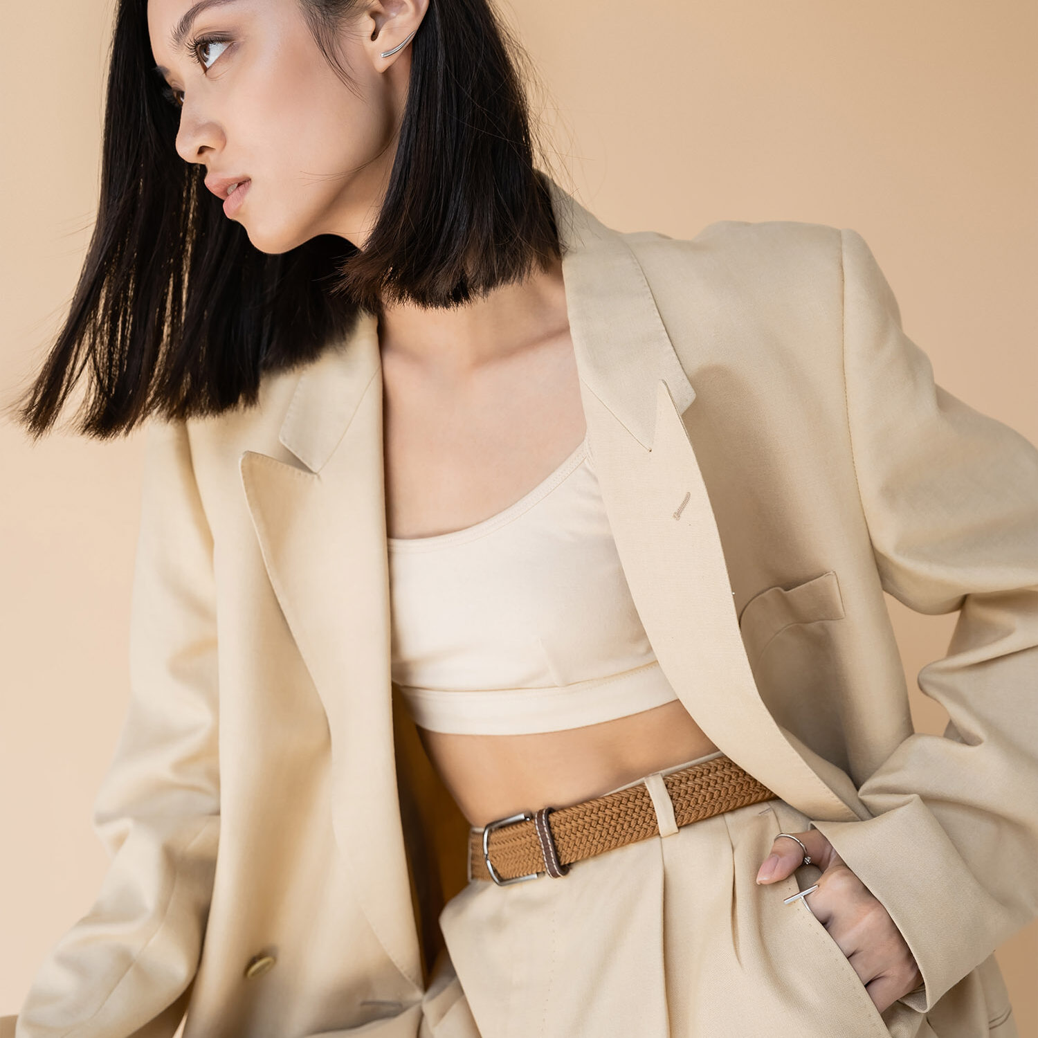 sensual asian woman in stylish pantsuit sitting in armchair and looking at camera on beige background